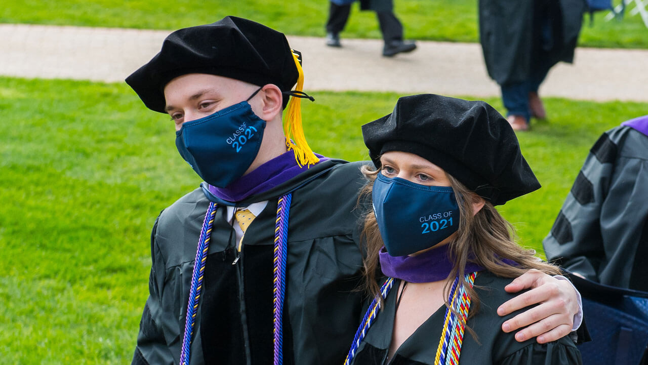 Two graduates walk with their arms around each other along the quad