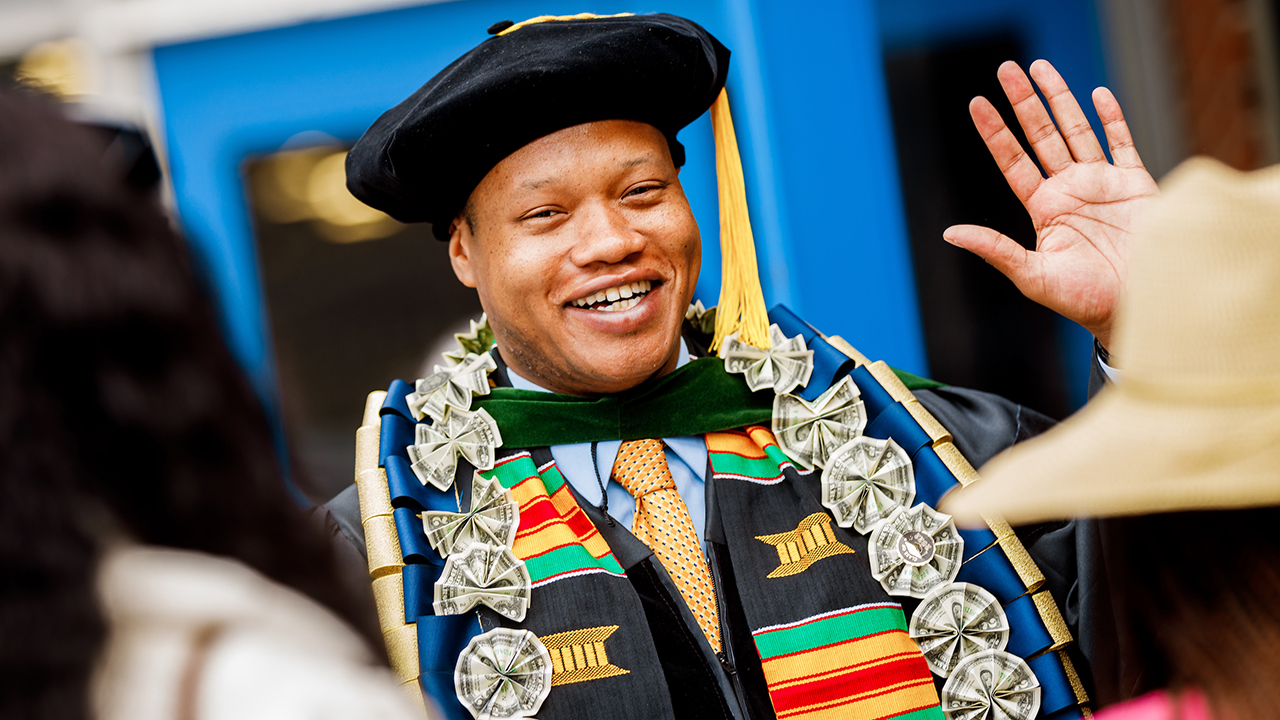 A graduate adorned with cords and stolls smiles and waves