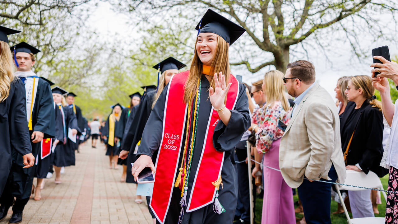 A graduate smiles and waves as she walks in a line of graduates