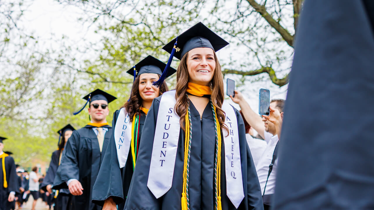 Two lines of students in caps and gowns walk down the quad path