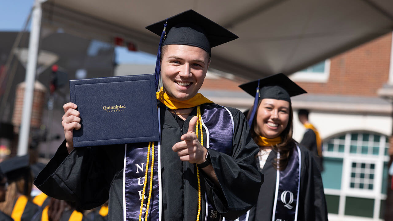 male graduate holds up his diploma and points at the camera