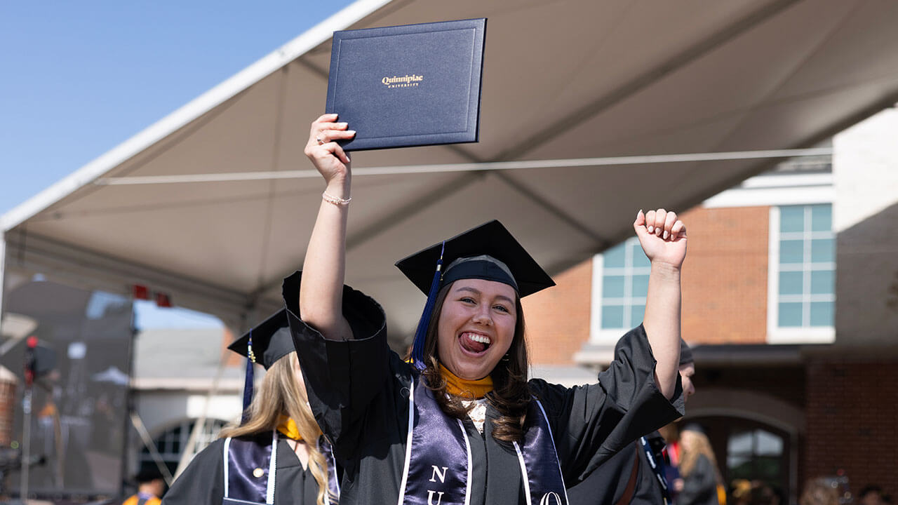 female graduate holds up her diploma and a raised fist while grinning broadly