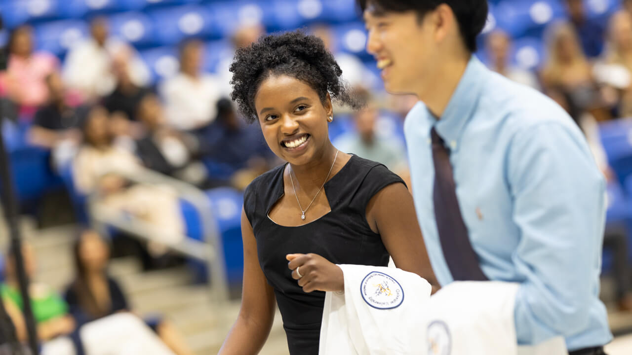 Two students smile as they walk into the ceremony with their white coats in their hands