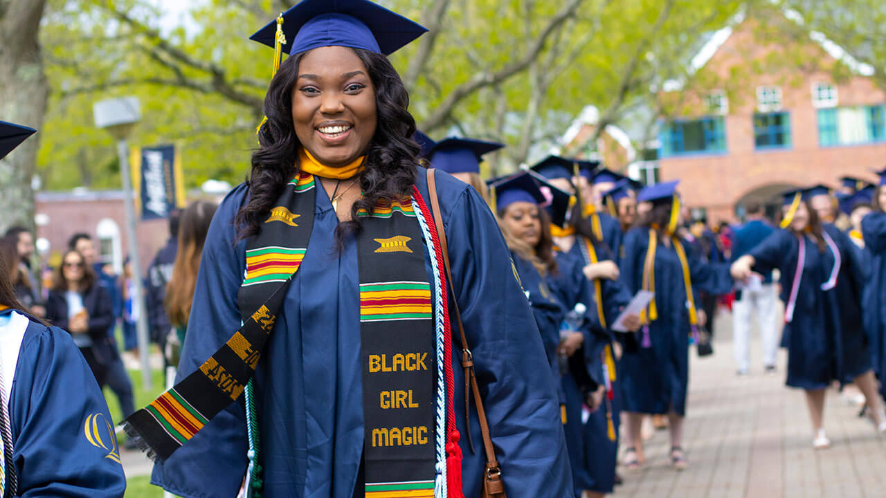 A graduate wearing a stole that reads "Black Girl Magic" walks down a brick path