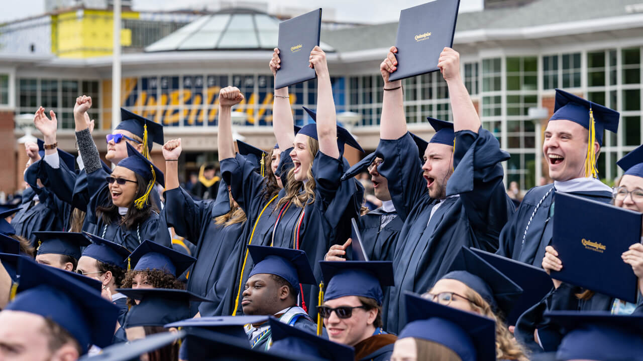 Graduates hold up their diplomas and cheer enthusiastically in front of the student center