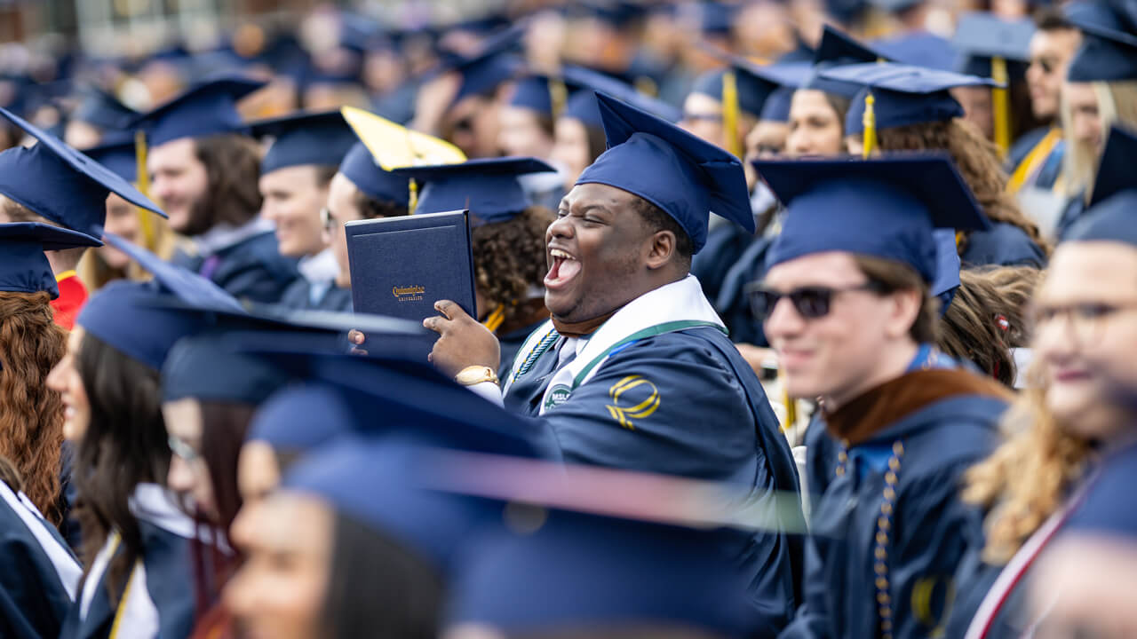 A graduate smiles hugely and shows his diploma to someone in the distance