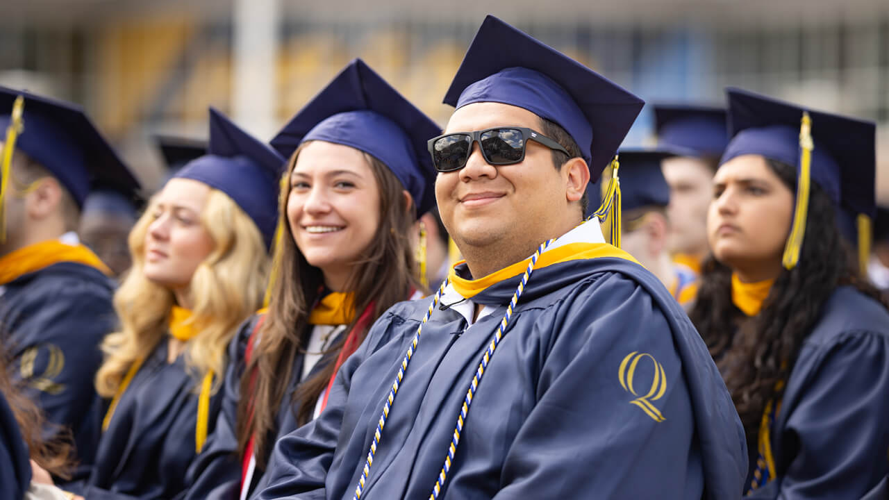 Several graduates sit in a row in their seats and smile