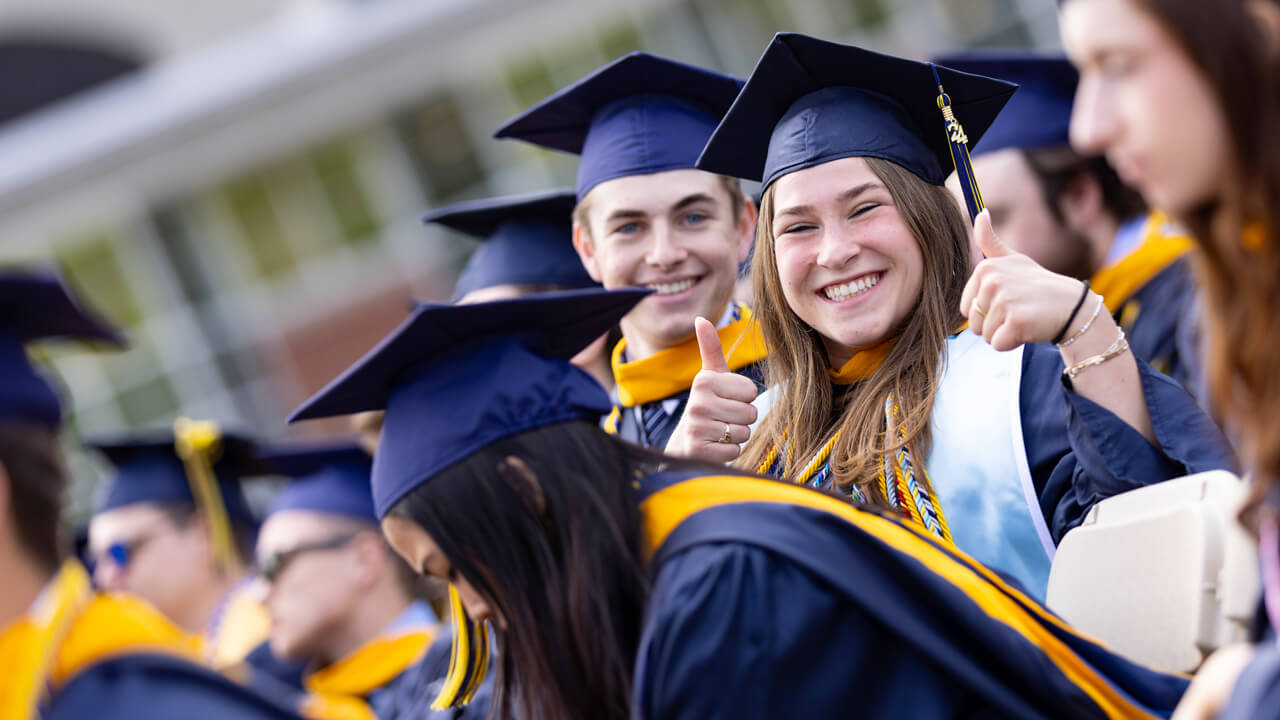 A graduate gives two thumbs up and smiles broadly from her seat