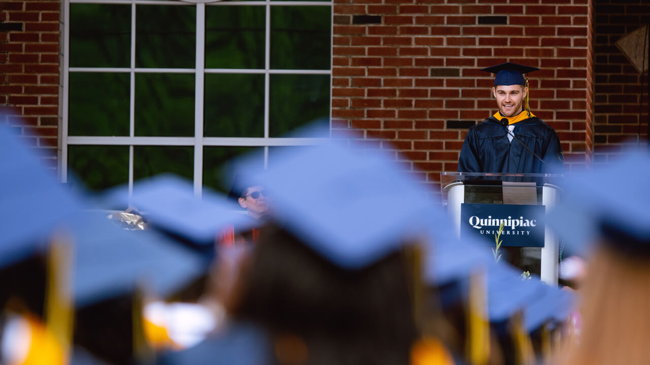 Alec Williams speaks on the library steps at a podium in front of dozens of graduates