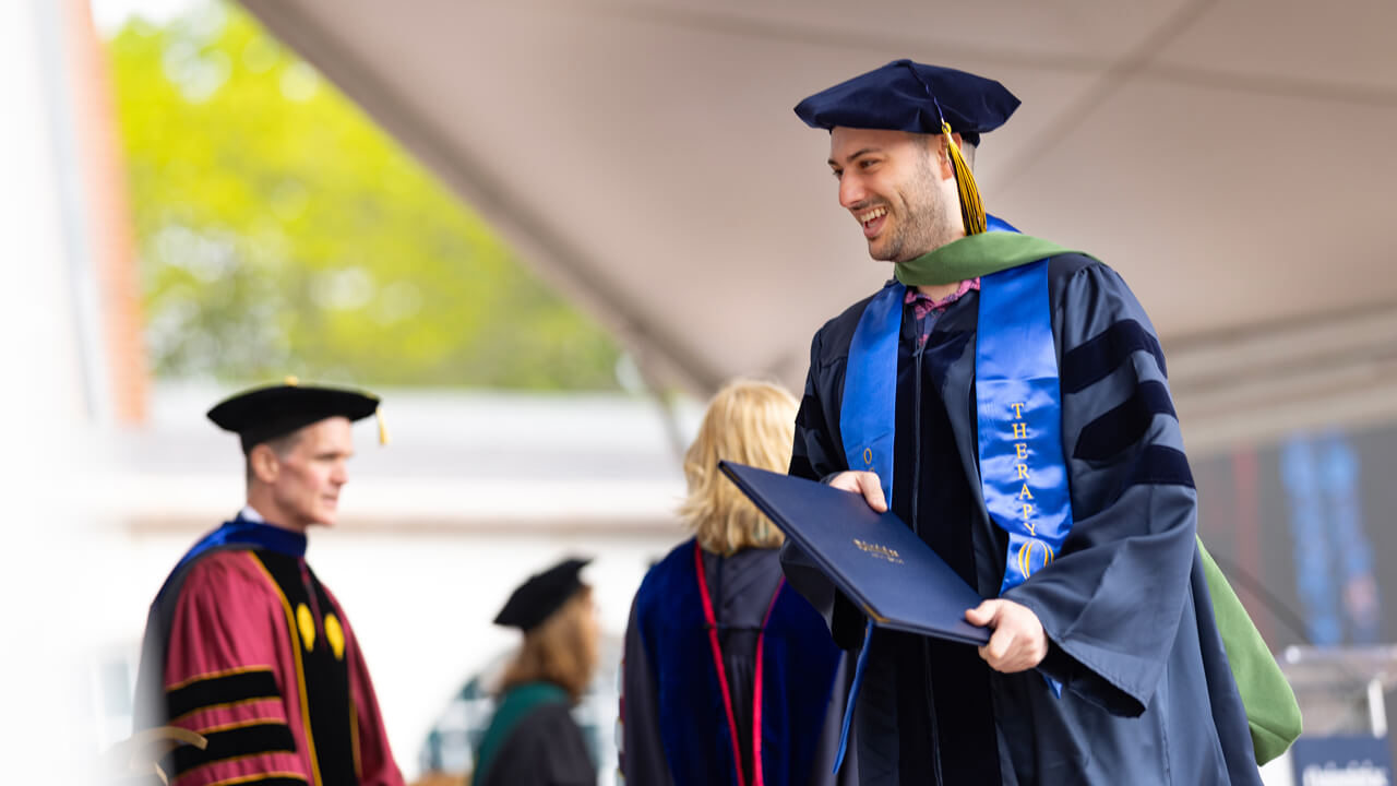 A graduate holds his diploma and smiles at a faculty member as he crosses the library steps