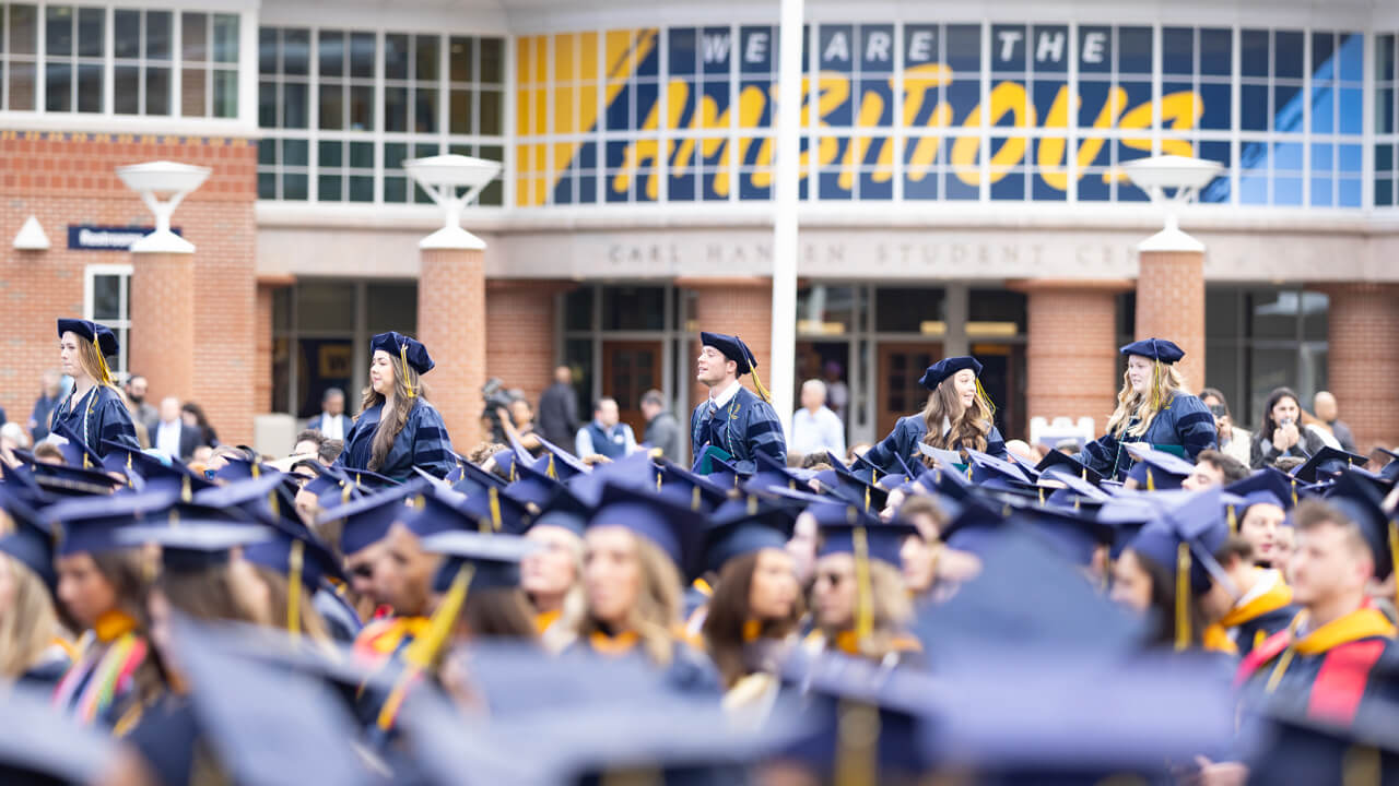 Dozens of graduates sit and some walk in front of the student center