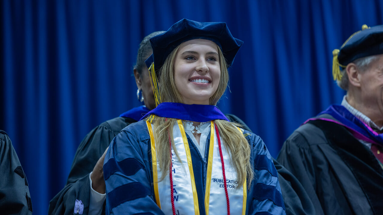 A graduate smiles proudly on stage while her wearing her doctoral hood and graduation stole and cords