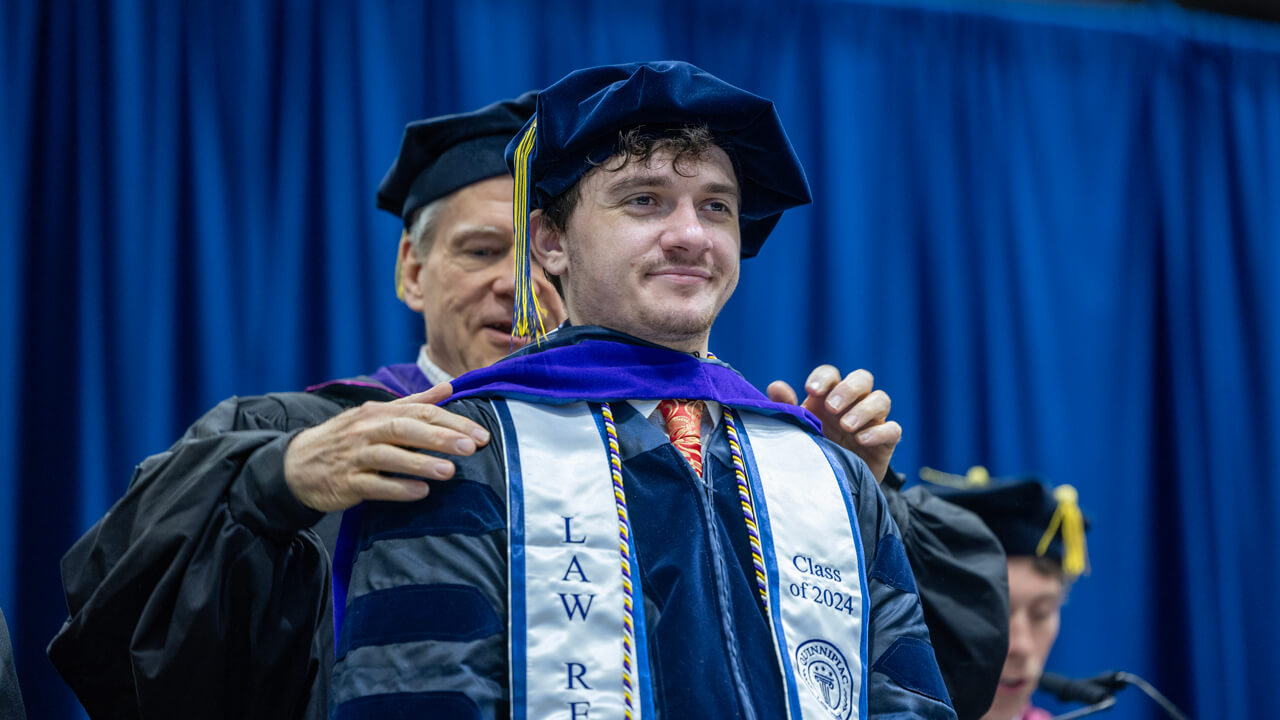 A professor places a doctoral hood on a graduate who wears a white stole that reads Class of 2024