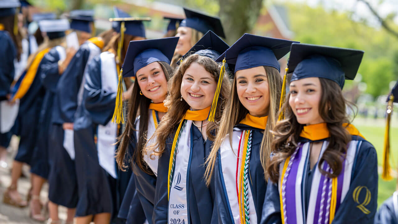 Four class of 2024 graduates smile broadly while waiting in line to take their seats.