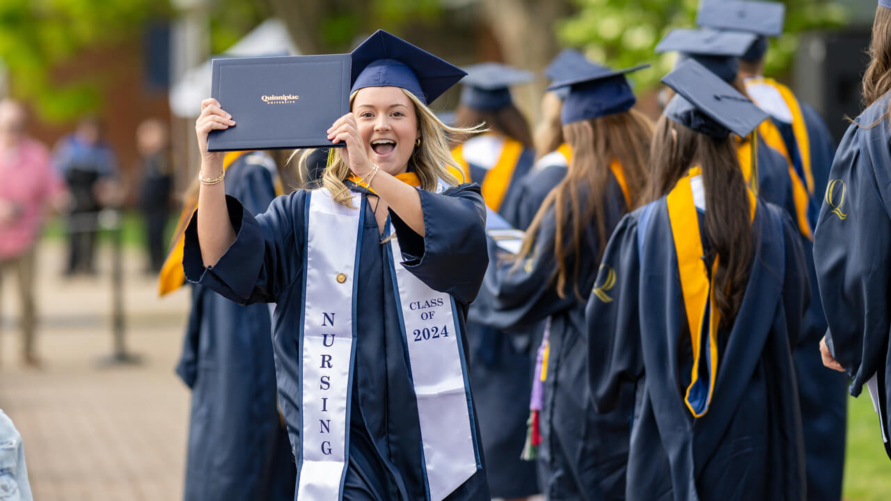 Graduate wearing a white stole that reads Nursing Class of 2024 holds up her diploma