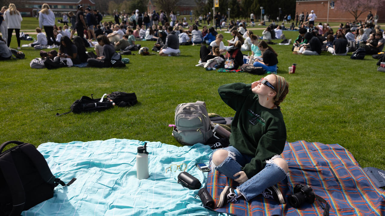 A student sitting on a blanket on the squad holds up solar eclipse glasses to their eyes.