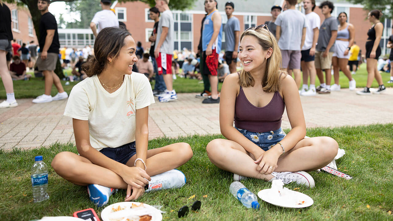 Two new students sit cross legged and smile together on the quad