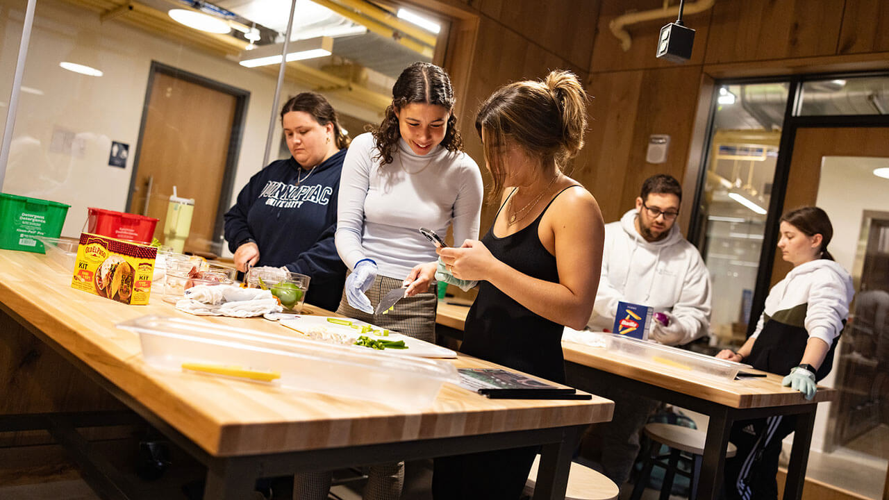 a female student in a black shirt takes a picture of the food in front of her while another students adds to her plate