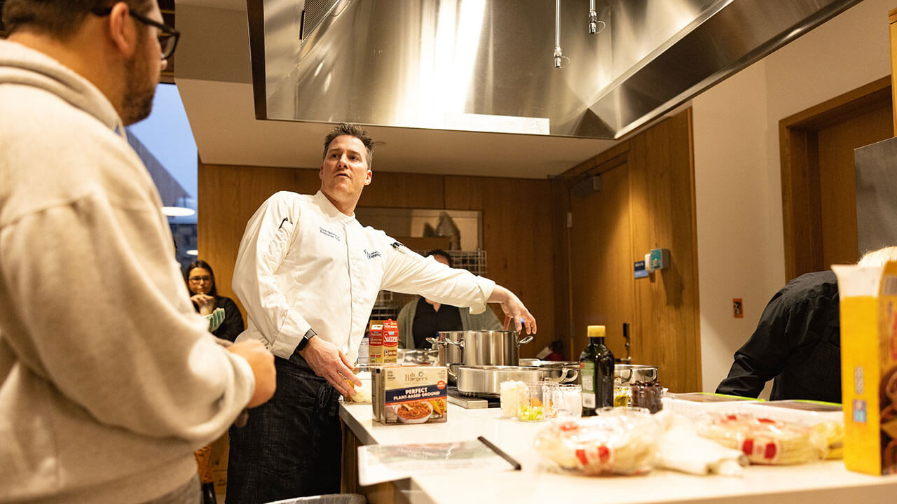 chef explains something to a male student, gesturing to the various pots and pans on the stove top