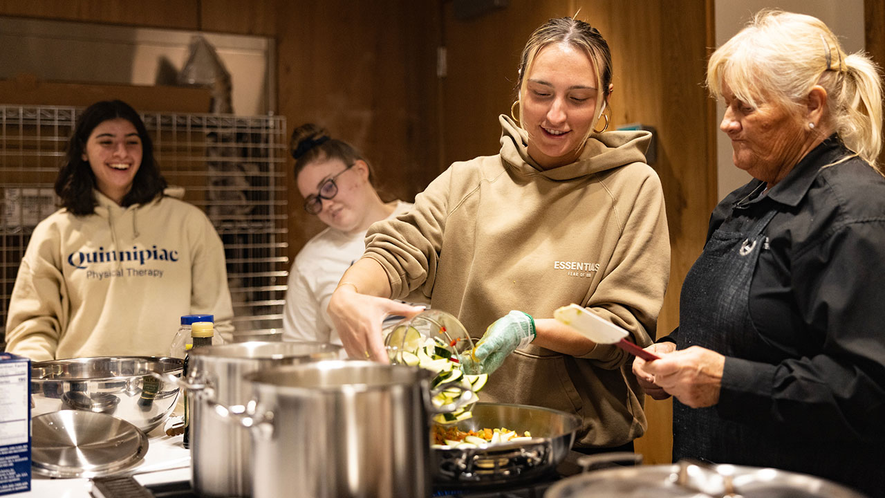 female student adds chopped vegetables to a skillet on the stove while a group of students watch