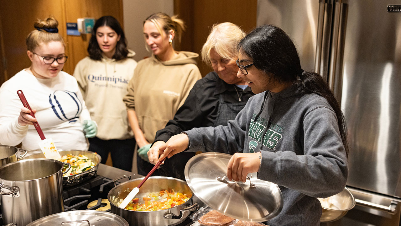 two students stir different skillets of vegetables while the assistant chef guides them