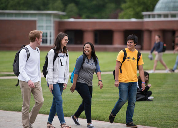 Four students in Quinnipiac shirts walk across the quad in front of the library