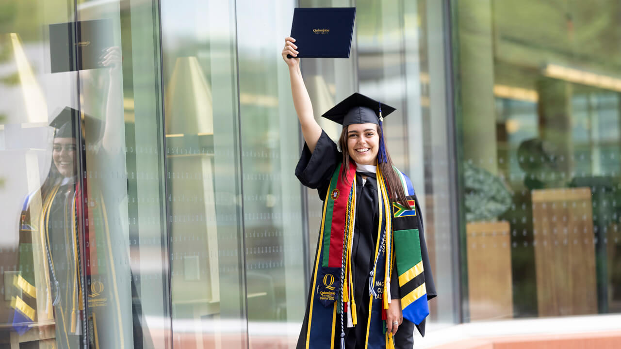 Gabriela Alvares wears her cap and gown and holds up her diploma in celebration outside the library