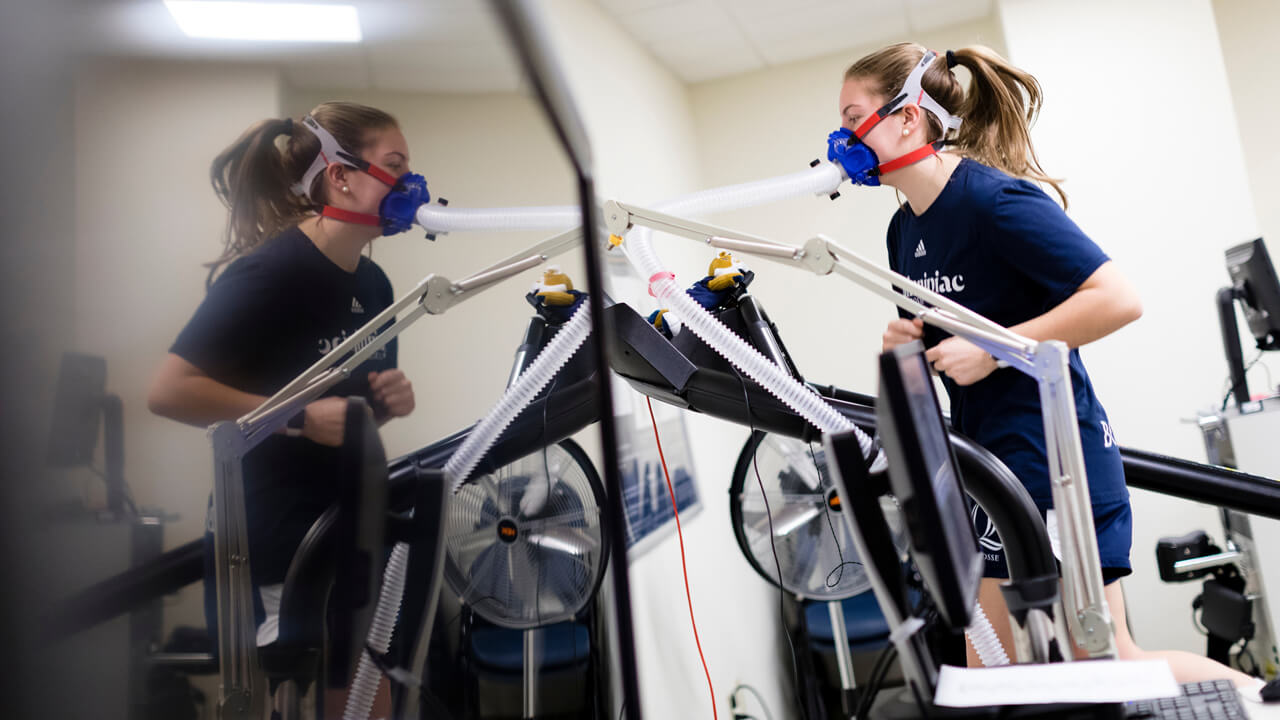 A student wears tracking devices as she runs on a treadmill with monitoring equipment around her