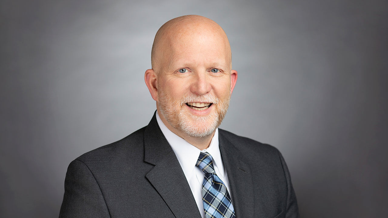 Headshot photo of John Bau wearing a black blazer against a gray backround.