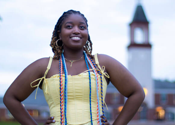 Student stands on quad with graduation cords in a yellow dress with the Arnold Bernard Library in the background