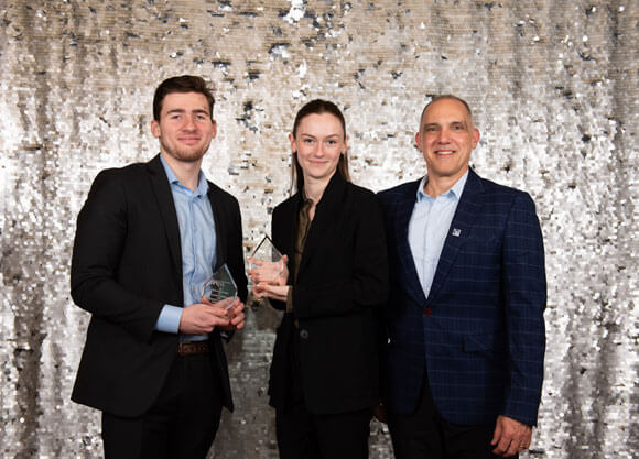 Two students and a professor stand in suits in front of a sequin backdrop holding award trophies.