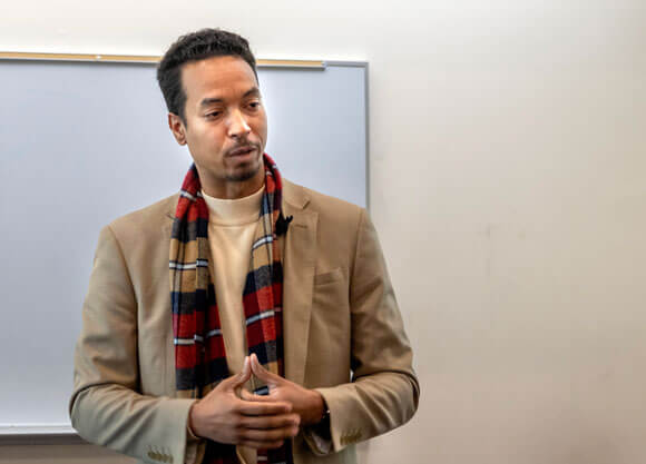 John Wood, wearing a scarf and jacket, speaks to a classroom.