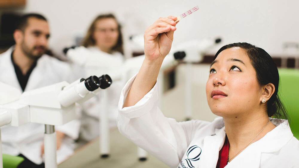 A student wearing a white lab coat holds a sample up to the light to inspect it