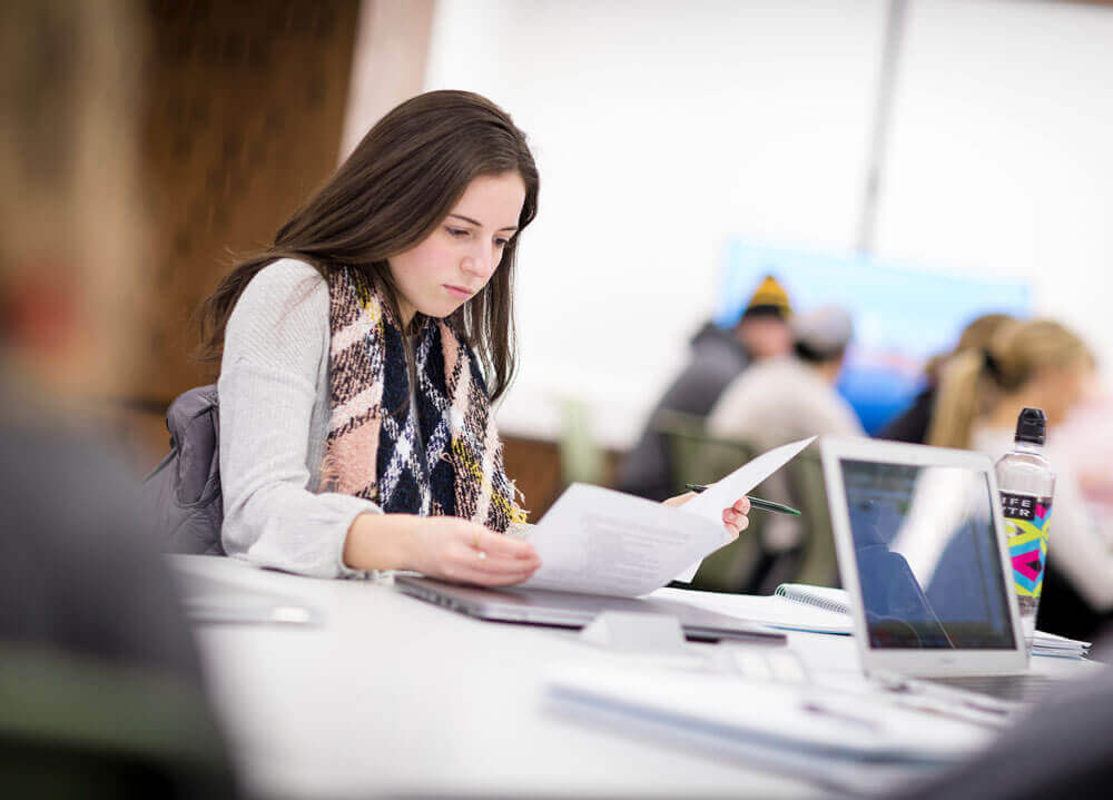 A student holding papers while sitting at a desk in a classroom