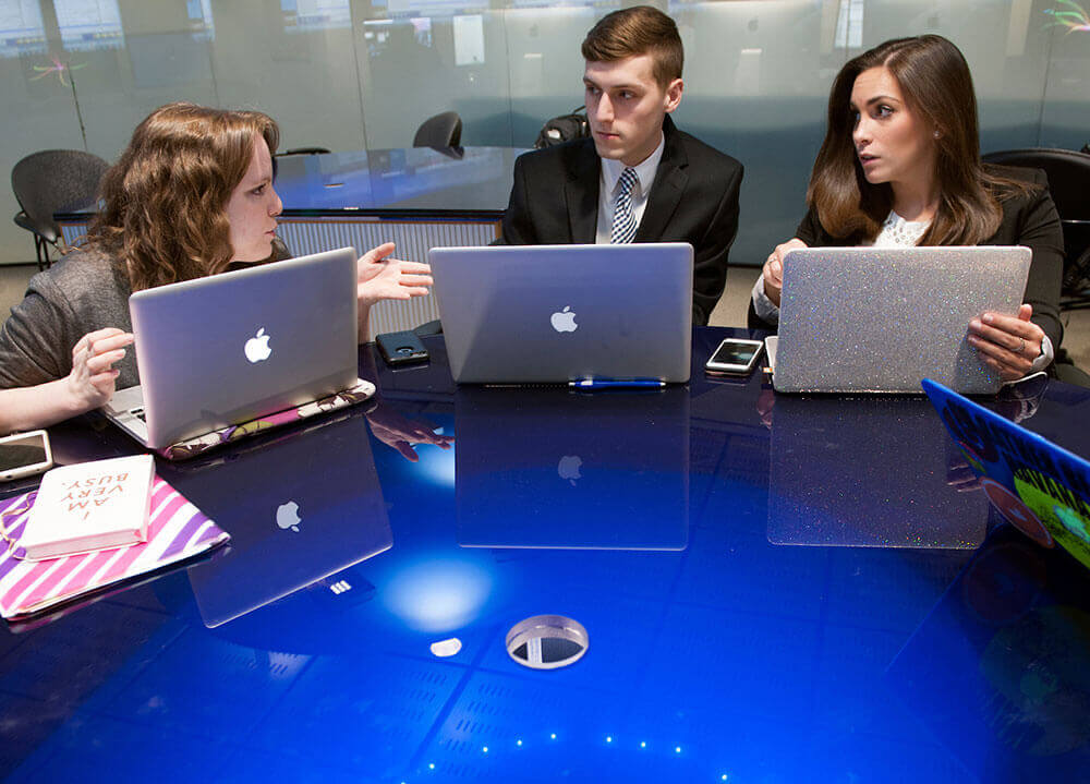 Three communications students sit side-by-side as they compare notes in preparation for a news course