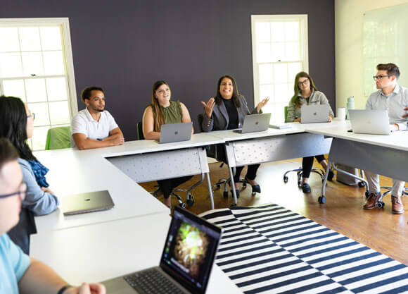 A group of adults sitting at a u-shaped table set up with laptops in front of them