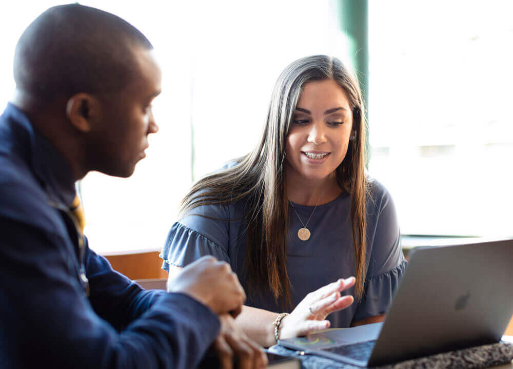 Two students sit at a table with a laptop in the lounge