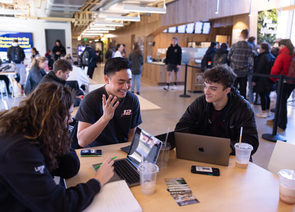 Students talk and work at their computers at a table near Shake Smart smoothie station