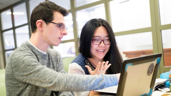 Two students sit at a table while looking at a laptop.