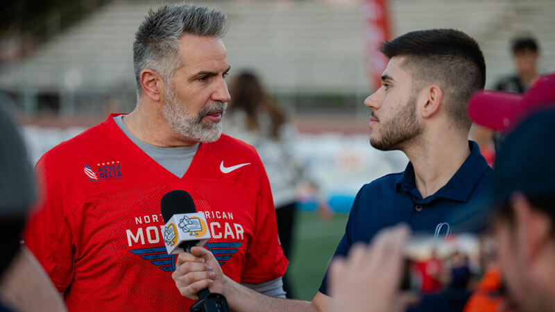 Santino Maione speaks with Kurt Warner on the sidelines of a football field